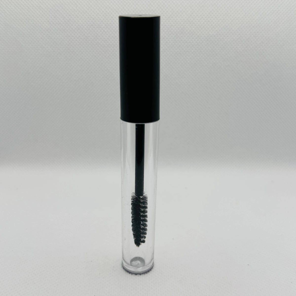 A clear cosmetic tube with a black cap and a black spoolie wand applicator inside, shown on a light gray textured background.