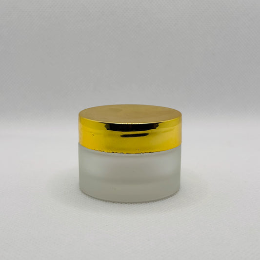 A small cosmetic jar with a frosted glass body and a shiny metallic gold lid, set against a neutral light gray background.