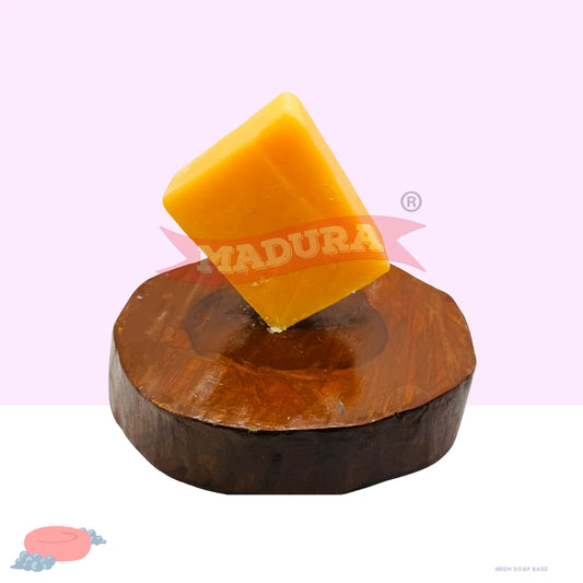 A rectangular bar of bright yellow soap standing upright on a round dark brown wooden base, photographed against a pale pink and white background. A red semi-transparent MADURA watermark overlays the center.